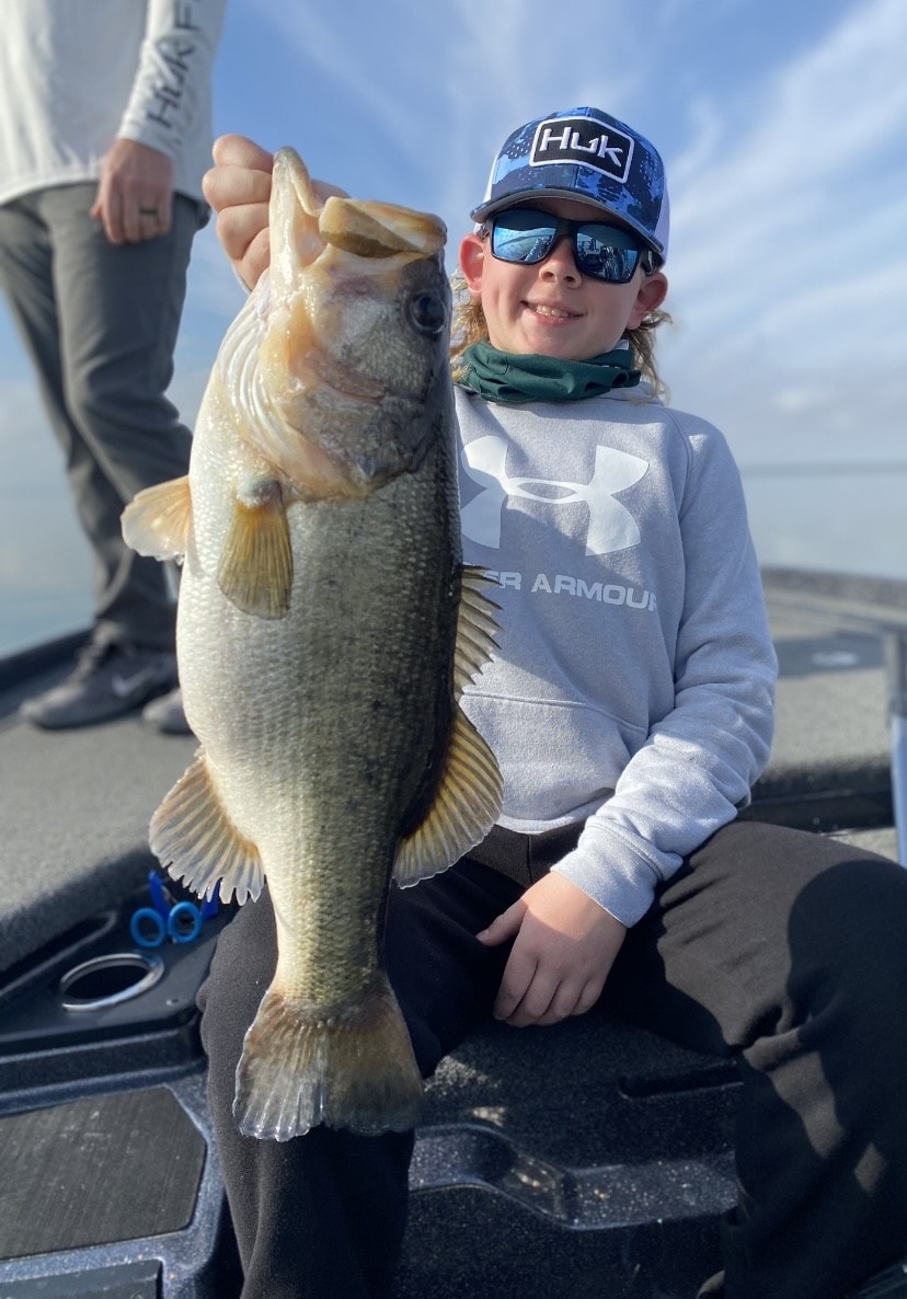 Kid in Huk cap and Under Armour hoodie holding big bass on sunny day
