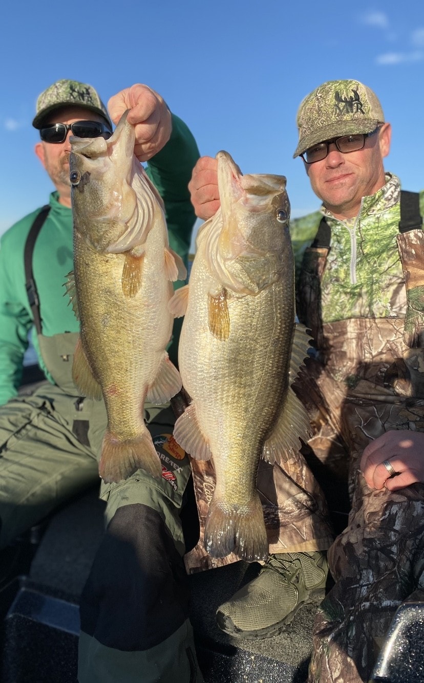 Two anglers in camo MR caps each holding big bass on sunny day