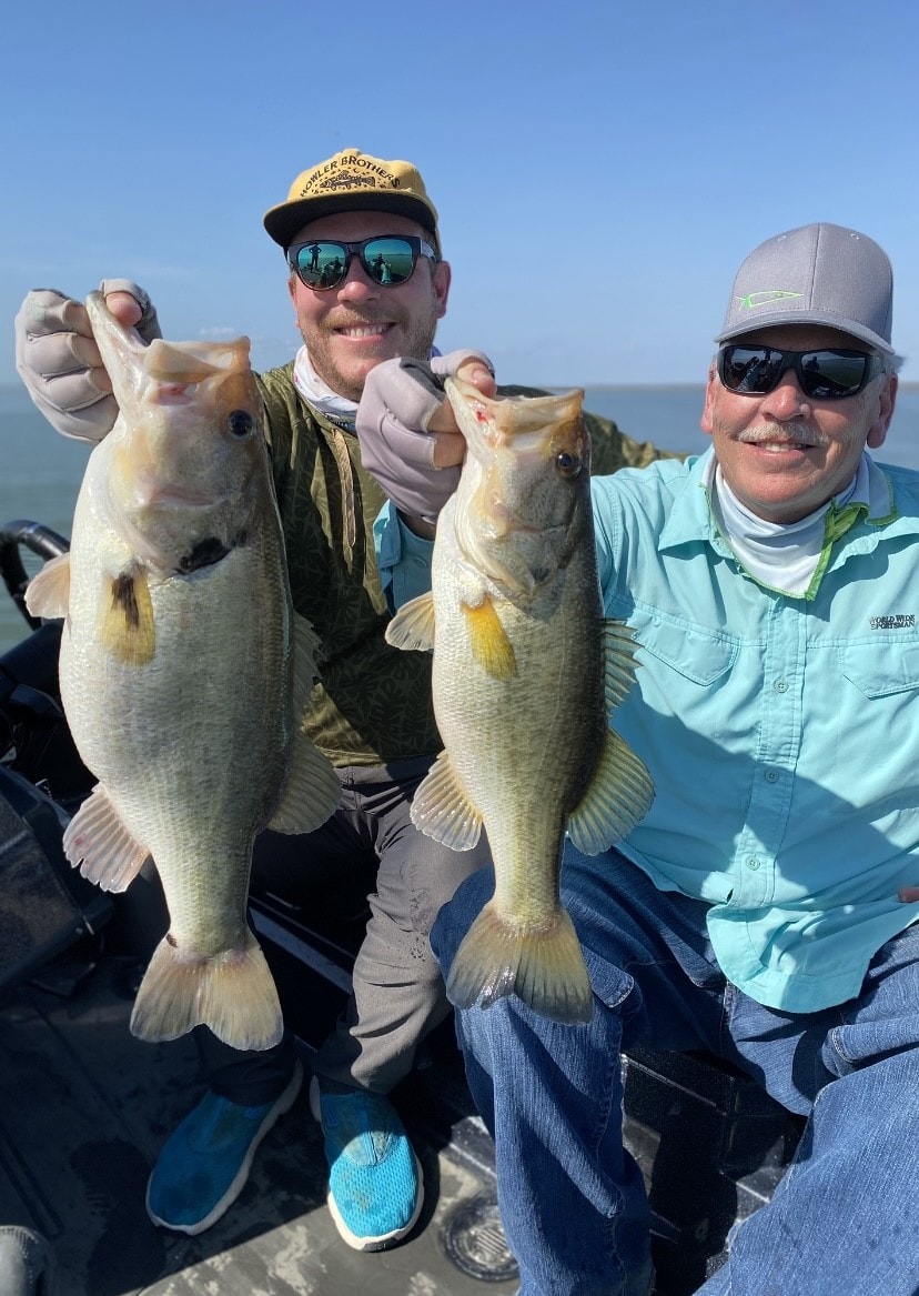 Two anglers in Howler Brothers and fishing caps each holding bass on sunny day