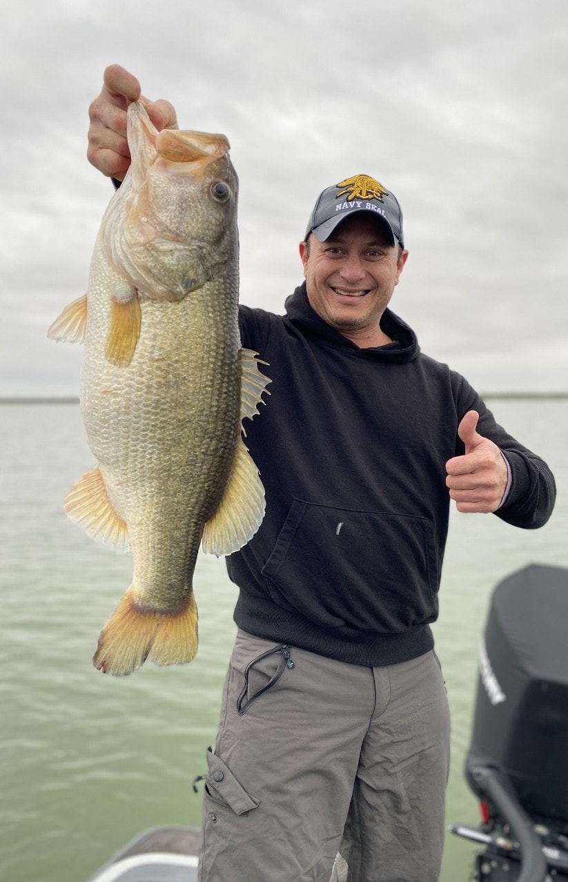 Man in Navy Seal cap holding big bass with thumbs up on cloudy day