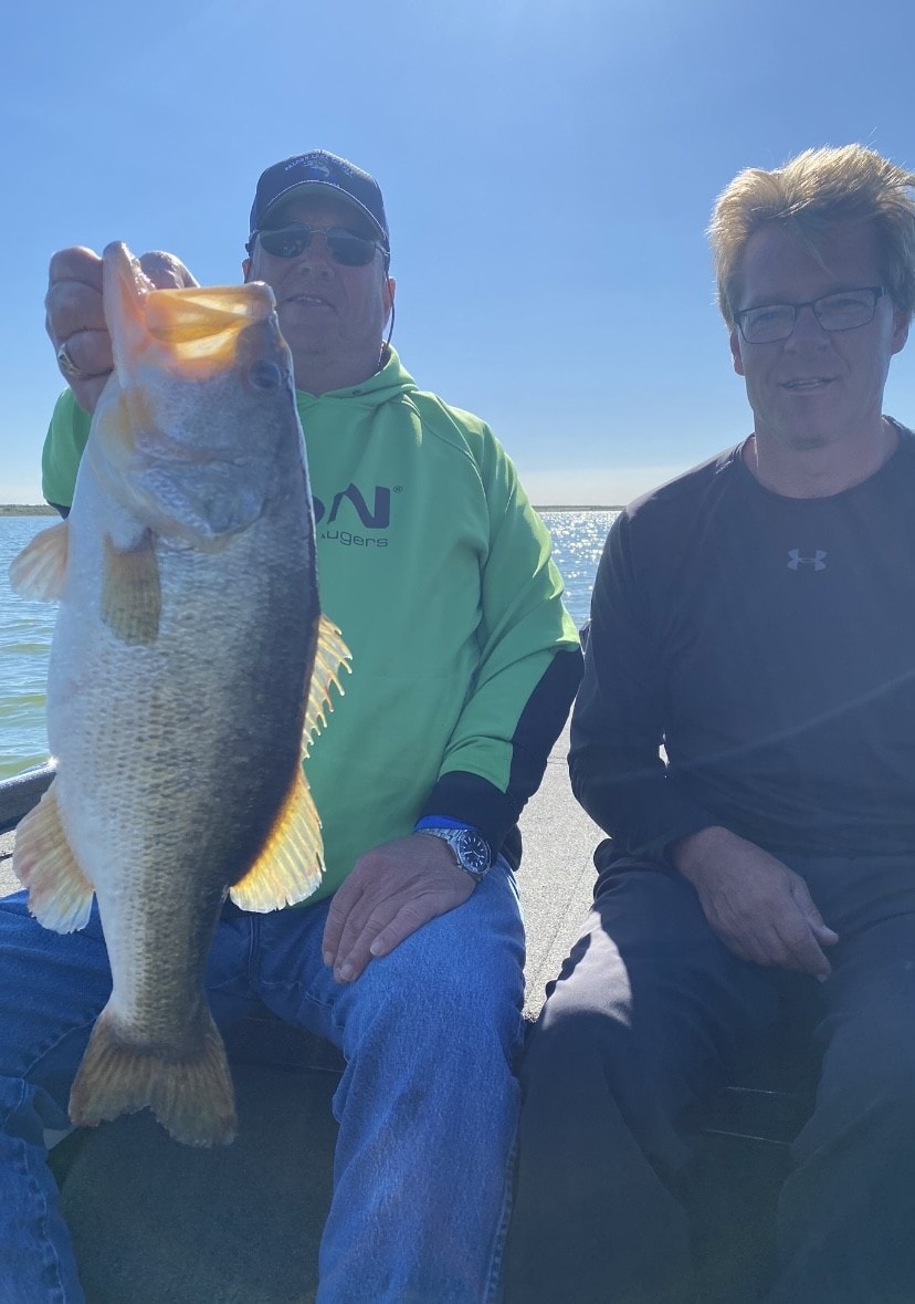 Two anglers on boat with man in green hoodie holding massive bass on sunny day