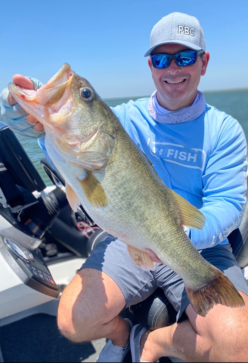 Man in PBC cap and Under Armour Fish shirt holding big bass on sunny day