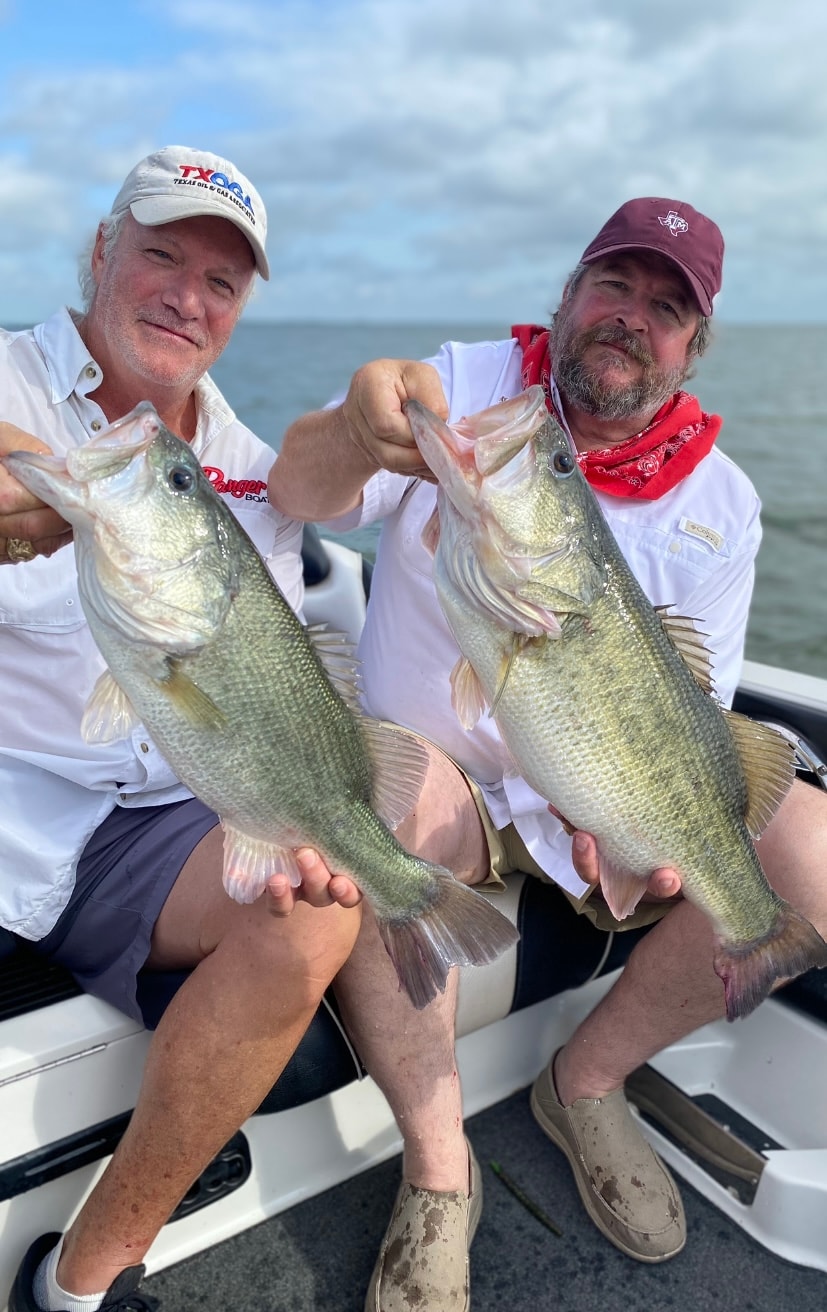 Two men in TXOGA and A&M caps each holding big bass on cloudy day