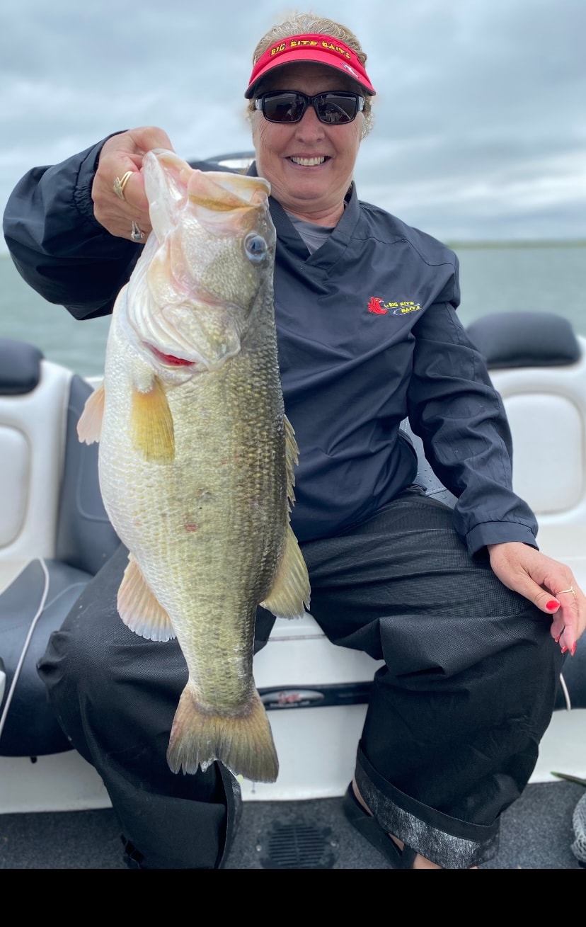 Woman in Big Bite Baits visor holding giant bass on cloudy day