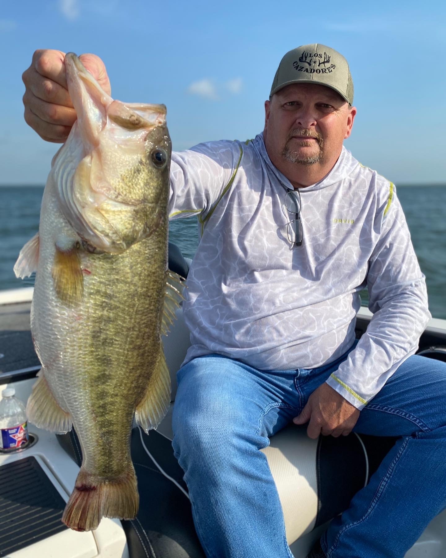 Man in Los Cazadores cap and Orvis shirt holding big bass on sunny day