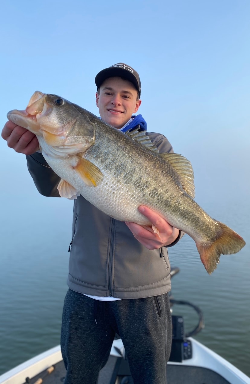 Teen in dark cap holding massive largemouth bass on foggy morning on Lake Falcon
