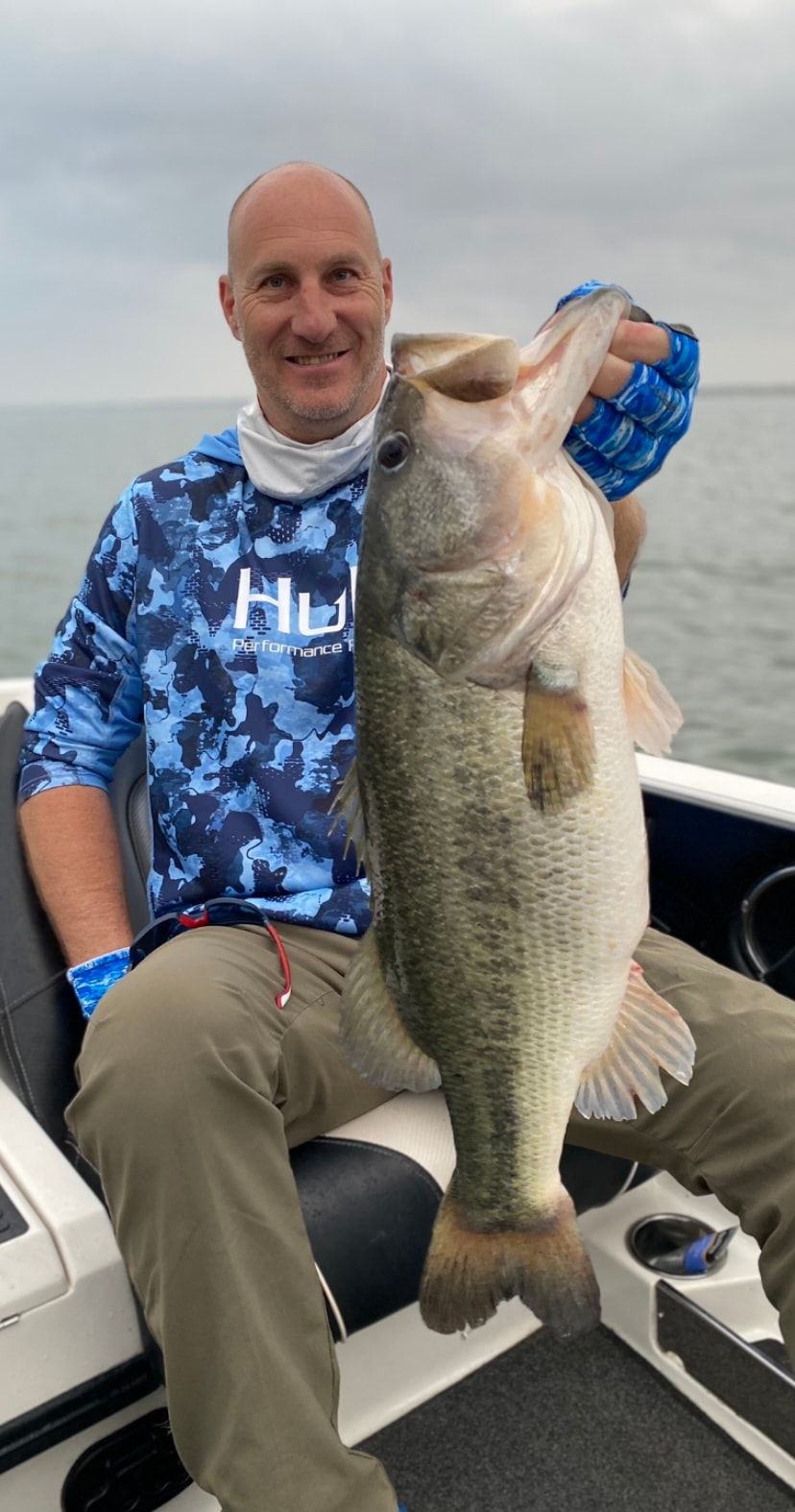 Bald man in Huk camo shirt holding massive bass on cloudy day on Lake Falcon