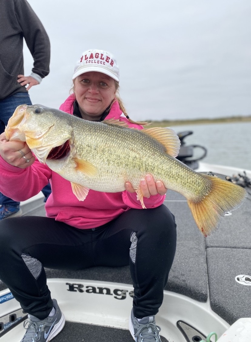 Woman in Flagler College cap and pink hoodie holding giant bass on cloudy day