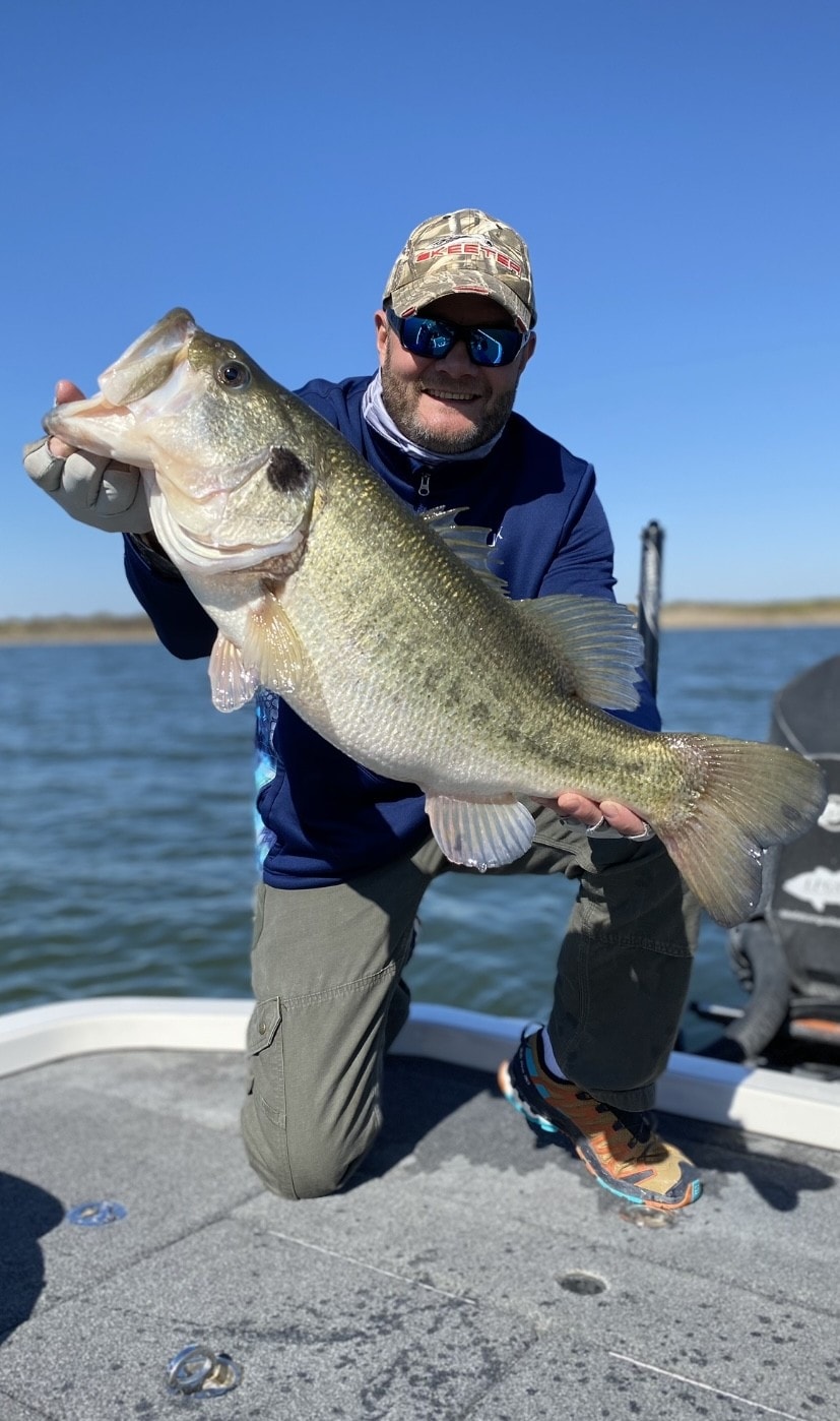 Man in Skeeter cap kneeling on boat holding massive bass on sunny day