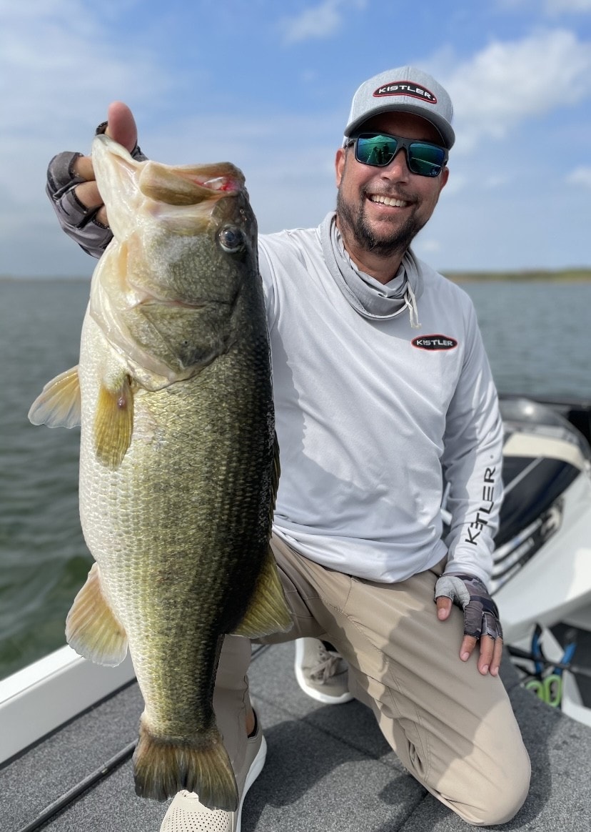 Man in Kistler shirt kneeling on boat holding large bass