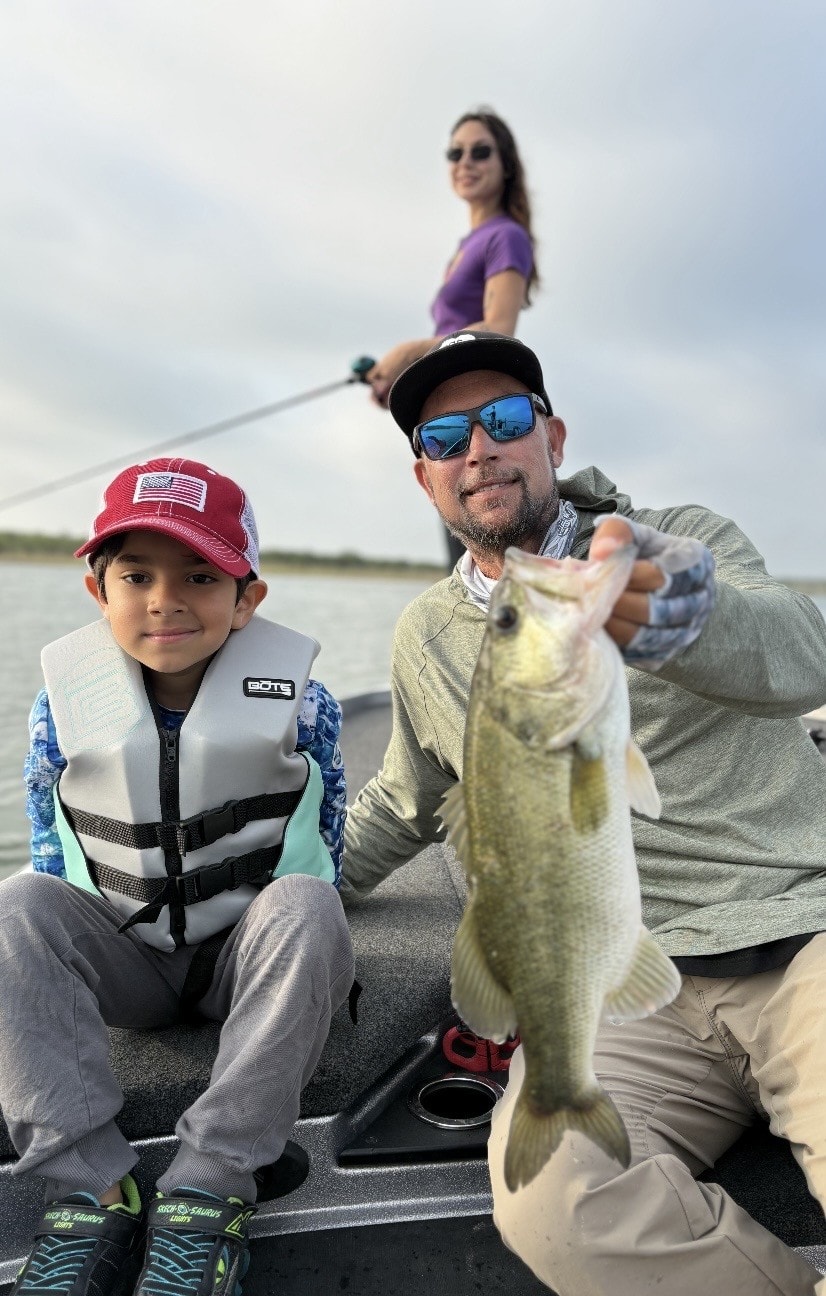 Man and young boy on boat holding bass with woman fishing in background