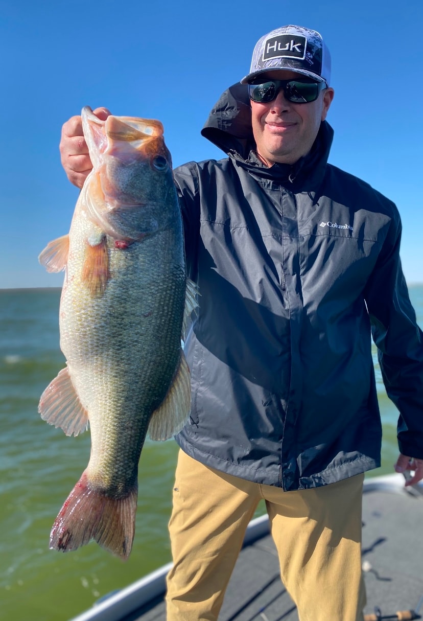Man in Huk cap and Columbia jacket holding trophy bass on sunny day