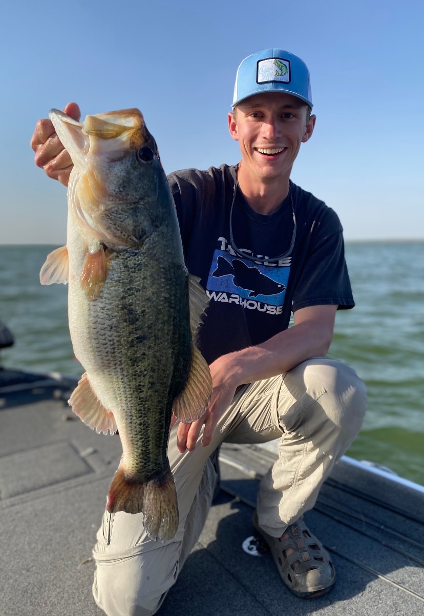 Young man in Tackle Warehouse shirt kneeling with big bass on sunny day