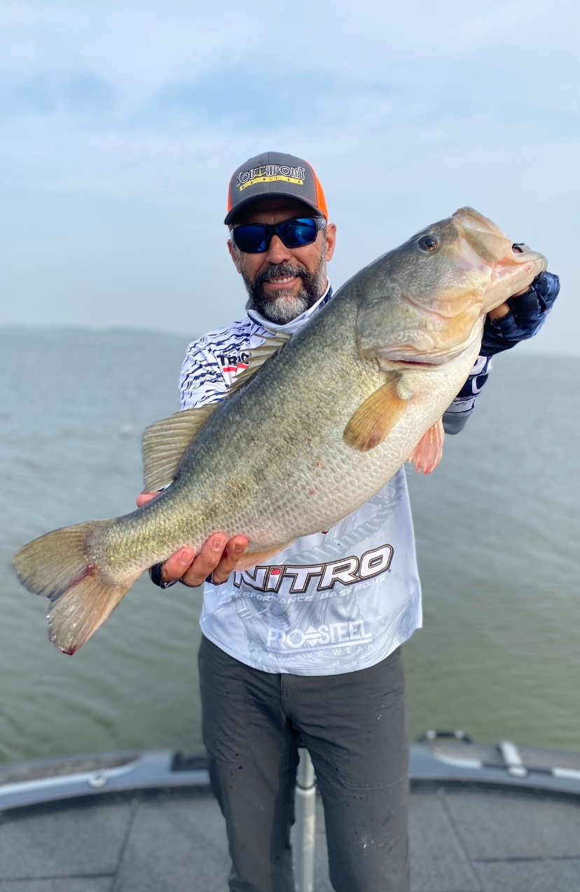 Man in Southpoint cap and Nitro shirt holding massive bass on cloudy day