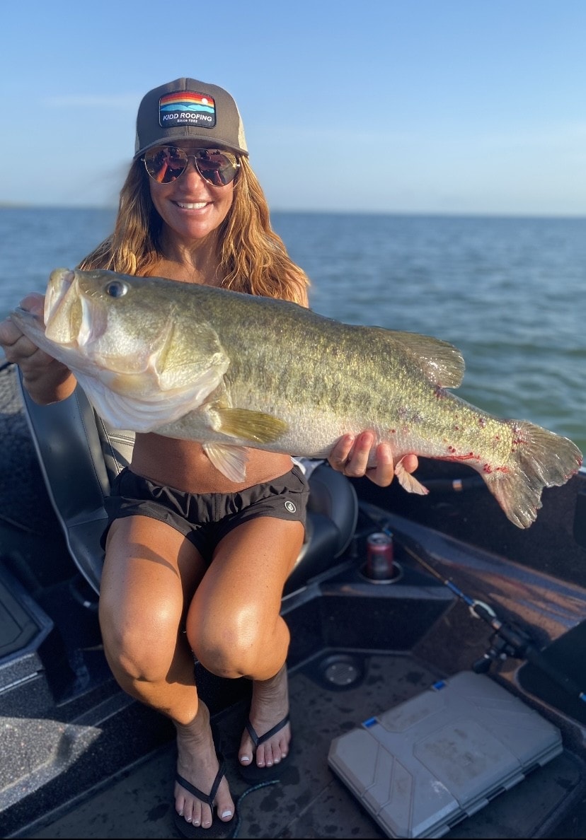 Woman in Kidd Roofing cap holding massive bass at golden hour