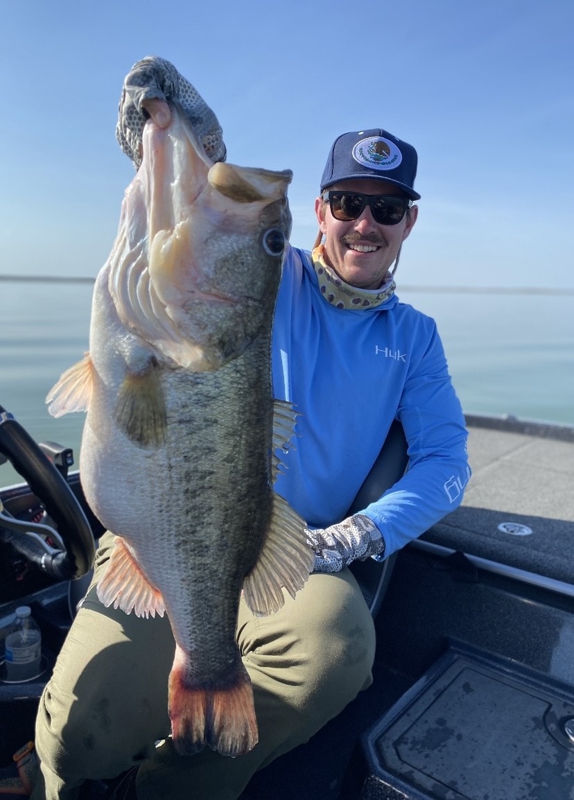 Man in Huk shirt holding massive bass upside down on sunny day