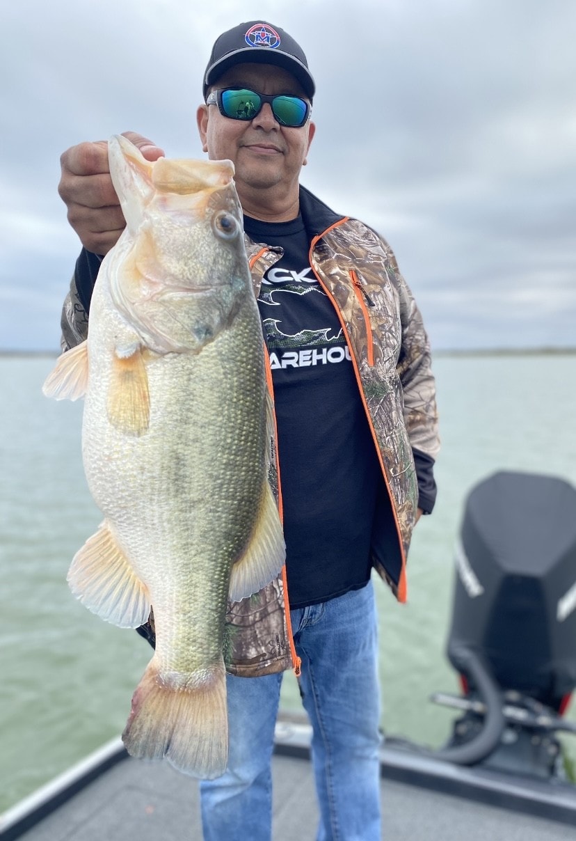 Man in camo jacket and Tackle Warehouse shirt holding big bass on cloudy day